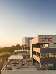 A modern architectural building with a geometric design is situated in a serene environment. The structure features multiple levels with large glass windows and balconies. In the foreground, a patio area with seating and a small covered section is visible. The area is well landscaped with trees and plants, and there is a small tram-like vehicle parked outside.