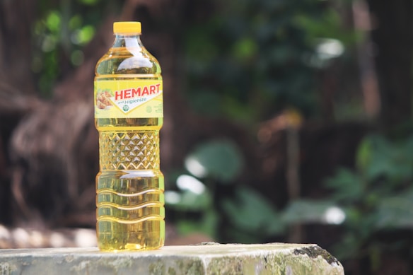 A plastic bottle filled with cooking oil is placed outdoors on a stone or concrete surface. The label on the bottle reads 'HEMART', with additional text. The background consists of out-of-focus greenery, including leaves and possibly tree trunks.