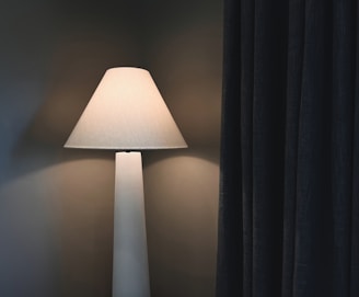 A warm light table lamp with a conical shade stands next to dark, textured curtains in a dimly lit room. The soft glow from the lamp casts a gentle shadow on the wall, highlighting the contrast between the light and the dark fabric of the curtains.
