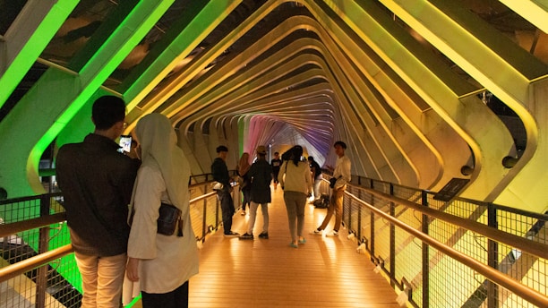 A vibrant pedestrian bridge with people walking across. The bridge is an architectural marvel featuring colorful LED lights in shades of green, yellow, and purple, creating a stunning visual effect. The structure's design includes crisscrossing beams and pathways, attracting several visitors who are taking photos and enjoying the aesthetics.