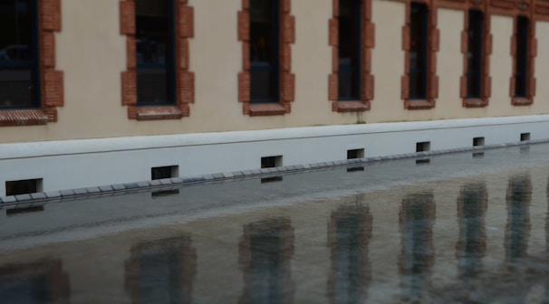 A facade of a building with multiple windows framed by brown bricks, reflected in a body of water. The wall is painted beige and features symmetrical window patterns.