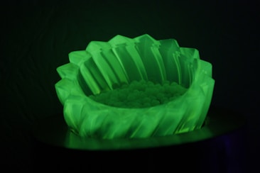 A glowing, translucent green bowl with a jagged rim, filled with small spherical beads. The image is dark, highlighting the luminescent quality of the bowl.