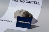 A piece of rough, textured rock placed on top of white paper with bold black text 'CASTRO CAPITAL'. In front of the rock, a navy blue card also with 'CASTRO CAPITAL' is displayed prominently.