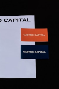 Two business cards placed on a white sheet. The cards are orange and dark blue, each with the text 'CASTRO CAPITAL' printed in uppercase.