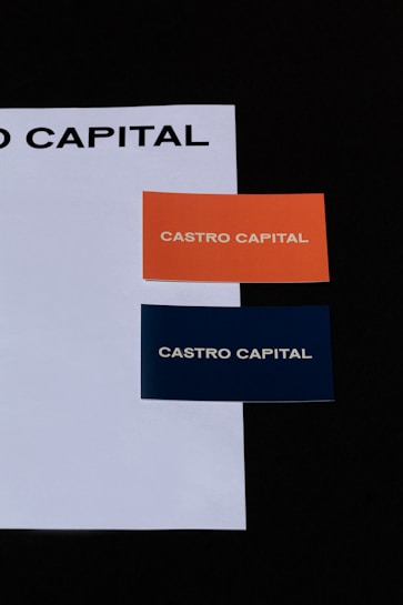 Two business cards placed on a white sheet. The cards are orange and dark blue, each with the text 'CASTRO CAPITAL' printed in uppercase.