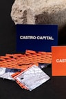 Several business cards are displayed in various colors and patterns. A dark blue card with white text reading 'Castro Capital' is prominently positioned in the background. In the foreground, there are numerous cards with woven red and orange designs, along with others featuring architectural imagery. Behind the blue card, a textured rock provides a contrasting natural element against the smooth cards.