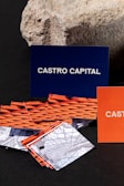 Several business cards are displayed in various colors and patterns. A dark blue card with white text reading 'Castro Capital' is prominently positioned in the background. In the foreground, there are numerous cards with woven red and orange designs, along with others featuring architectural imagery. Behind the blue card, a textured rock provides a contrasting natural element against the smooth cards.