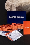 Several business cards are displayed in various colors and patterns. A dark blue card with white text reading 'Castro Capital' is prominently positioned in the background. In the foreground, there are numerous cards with woven red and orange designs, along with others featuring architectural imagery. Behind the blue card, a textured rock provides a contrasting natural element against the smooth cards.
