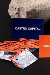 Several business cards are displayed in various colors and patterns. A dark blue card with white text reading 'Castro Capital' is prominently positioned in the background. In the foreground, there are numerous cards with woven red and orange designs, along with others featuring architectural imagery. Behind the blue card, a textured rock provides a contrasting natural element against the smooth cards.