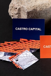 Several business cards are displayed in various colors and patterns. A dark blue card with white text reading 'Castro Capital' is prominently positioned in the background. In the foreground, there are numerous cards with woven red and orange designs, along with others featuring architectural imagery. Behind the blue card, a textured rock provides a contrasting natural element against the smooth cards.