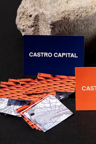 Several business cards are displayed in various colors and patterns. A dark blue card with white text reading 'Castro Capital' is prominently positioned in the background. In the foreground, there are numerous cards with woven red and orange designs, along with others featuring architectural imagery. Behind the blue card, a textured rock provides a contrasting natural element against the smooth cards.