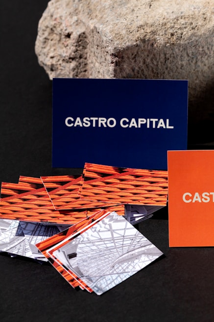 Several business cards are displayed in various colors and patterns. A dark blue card with white text reading 'Castro Capital' is prominently positioned in the background. In the foreground, there are numerous cards with woven red and orange designs, along with others featuring architectural imagery. Behind the blue card, a textured rock provides a contrasting natural element against the smooth cards.