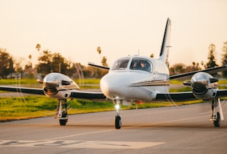 A small twin-engine airplane is on a runway with propellers in motion. The aircraft is sleek and white, with a person visible through the cockpit window. The background shows a lush green field and several palm trees under a warm, late afternoon sky.