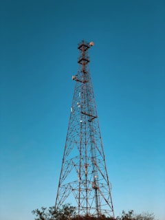 A tall metal lattice telecommunications tower stands against a clear blue sky. The tower is equipped with various antennas and dishes, indicating its function for signal transmission. Nearby, the tops of trees are visible, which suggests the tower's location in an open area or a field.