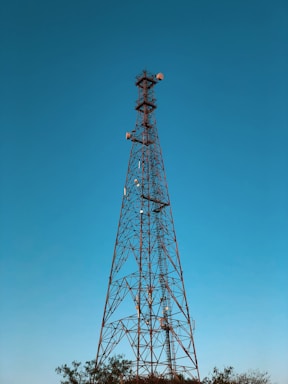 A tall metal lattice telecommunications tower stands against a clear blue sky. The tower is equipped with various antennas and dishes, indicating its function for signal transmission. Nearby, the tops of trees are visible, which suggests the tower's location in an open area or a field.