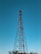 A tall metal lattice telecommunications tower stands against a clear blue sky. The tower is equipped with various antennas and dishes, indicating its function for signal transmission. Nearby, the tops of trees are visible, which suggests the tower's location in an open area or a field.