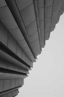 An architectural structure composed of layered, metallic panels that seem to fan outwards, creating a sense of depth and perspective. The panels have a textured, perforated appearance and the overall composition conveys a modern and geometric aesthetic.