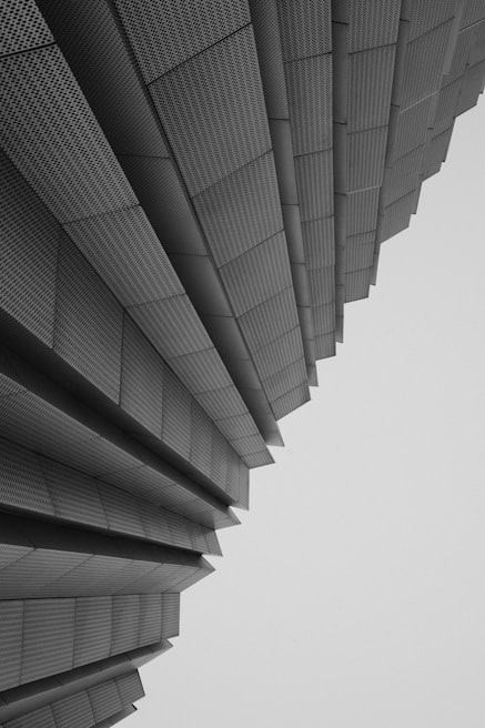 An architectural structure composed of layered, metallic panels that seem to fan outwards, creating a sense of depth and perspective. The panels have a textured, perforated appearance and the overall composition conveys a modern and geometric aesthetic.