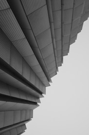 An architectural structure composed of layered, metallic panels that seem to fan outwards, creating a sense of depth and perspective. The panels have a textured, perforated appearance and the overall composition conveys a modern and geometric aesthetic.