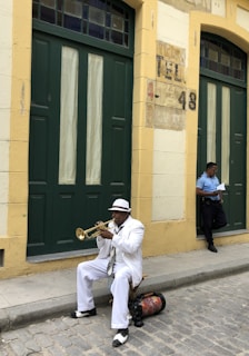 A man dressed in a white suit and hat sits on a small stool on a cobblestone street, playing a trumpet. He is positioned in front of a building with large, dark green doors and yellow walls. To the right, another man in a blue uniform leans against the building, looking at a piece of paper.