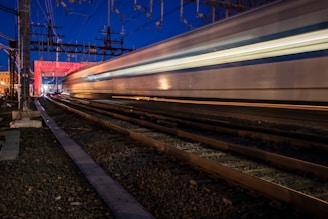 A high-speed train moves swiftly along tracks, capturing motion blur against a backdrop of nighttime sky and industrial structures. The lights on the train create streaks of illumination, reflecting the speed and dynamic energy of the scene. Power lines and infrastructure are prominent above the tracks.