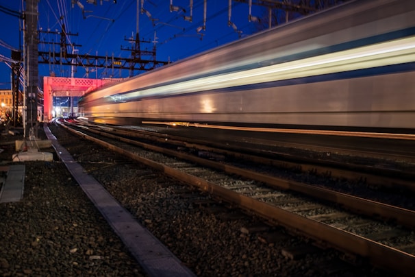 A high-speed train moves swiftly along tracks, capturing motion blur against a backdrop of nighttime sky and industrial structures. The lights on the train create streaks of illumination, reflecting the speed and dynamic energy of the scene. Power lines and infrastructure are prominent above the tracks.