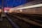 A high-speed train moves swiftly along tracks, capturing motion blur against a backdrop of nighttime sky and industrial structures. The lights on the train create streaks of illumination, reflecting the speed and dynamic energy of the scene. Power lines and infrastructure are prominent above the tracks.