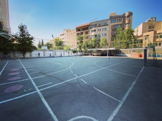 An outdoor sports court surrounded by tall buildings and trees, featuring a basketball hoop and a volleyball net. The court has white lines marking the different sections on the asphalt surface. The architecture in the background includes modern residential buildings with large windows.