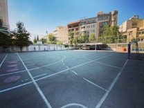 An outdoor sports court surrounded by tall buildings and trees, featuring a basketball hoop and a volleyball net. The court has white lines marking the different sections on the asphalt surface. The architecture in the background includes modern residential buildings with large windows.