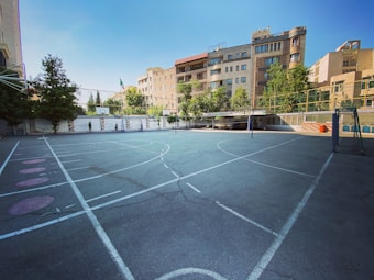 An outdoor sports court surrounded by tall buildings and trees, featuring a basketball hoop and a volleyball net. The court has white lines marking the different sections on the asphalt surface. The architecture in the background includes modern residential buildings with large windows.