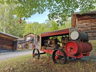 An old, red and rusted farm machine with large wheels is situated in a rustic setting surrounded by wooden buildings and greenery. The machine appears to be an antique agricultural equipment, possibly a tractor or engine. Overhead, green leaves from a tree branch create a natural canopy.