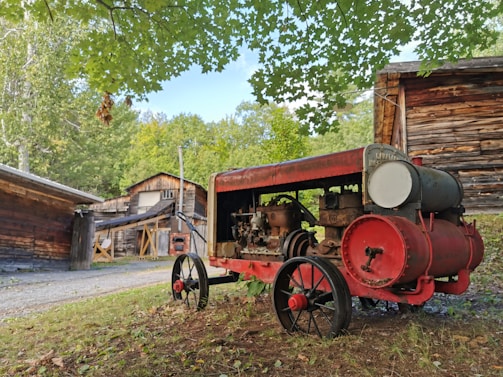 An old, red and rusted farm machine with large wheels is situated in a rustic setting surrounded by wooden buildings and greenery. The machine appears to be an antique agricultural equipment, possibly a tractor or engine. Overhead, green leaves from a tree branch create a natural canopy.