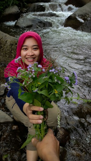 A person wearing a bright red headscarf smiles warmly while holding out a bouquet of wildflowers towards the camera. In the background, a small stream flows over rocks, creating a serene natural setting.