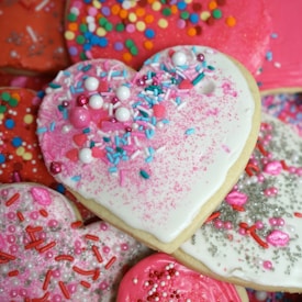 Heart-shaped cookies with vibrant icing and colorful sprinkles are artfully arranged. The icing is predominantly white and pink, with a variety of sprinkles including round pearls, tiny hearts, and jimmies in multiple colors.