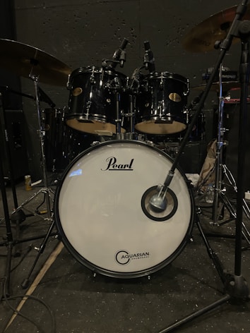 A black drum set is prominently displayed, featuring a large bass drum with the brand logos 'Pearl' and 'Aquarian Drumheads' visible on the front. Microphones are positioned above the drums, possibly for recording or amplification, and cymbals are partially visible. The setup is in a dimly lit room, likely a practice or recording space.