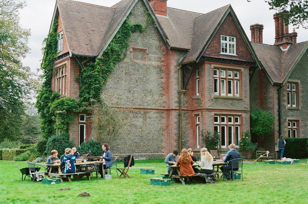 Un grupo de personas sentadas en mesas de picnic frente a un gran edificio de ladrillo