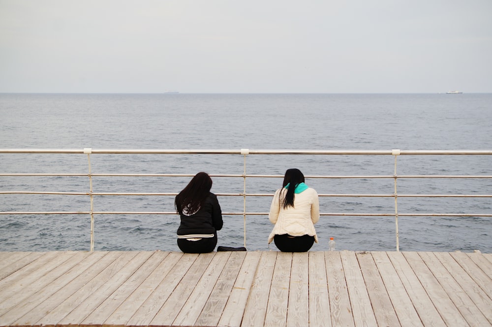 two women sitting on a pier looking out at the ocean
