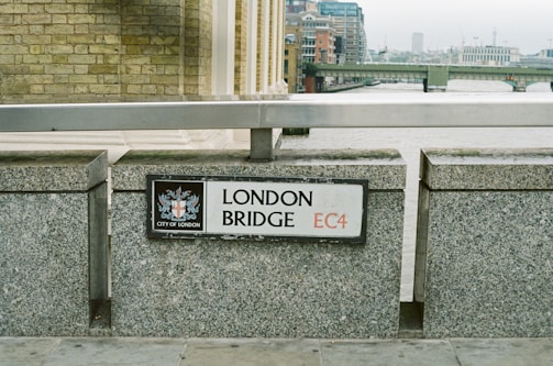 A street sign reading 'London Bridge EC4' is mounted on a stone wall. The sign includes the crest of the City of London. In the background, an urban landscape with a river and multiple bridges can be seen on an overcast day.