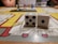 Two white dice with black dots are placed on a colorful board game. The board shows various rooms and paths outlined in red, yellow, and gray. In the background, there are blurred figures and a few plastic game pieces in red, blue, and yellow.