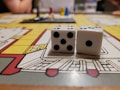 Two white dice with black dots are placed on a colorful board game. The board shows various rooms and paths outlined in red, yellow, and gray. In the background, there are blurred figures and a few plastic game pieces in red, blue, and yellow.