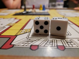 Two white dice with black dots are placed on a colorful board game. The board shows various rooms and paths outlined in red, yellow, and gray. In the background, there are blurred figures and a few plastic game pieces in red, blue, and yellow.