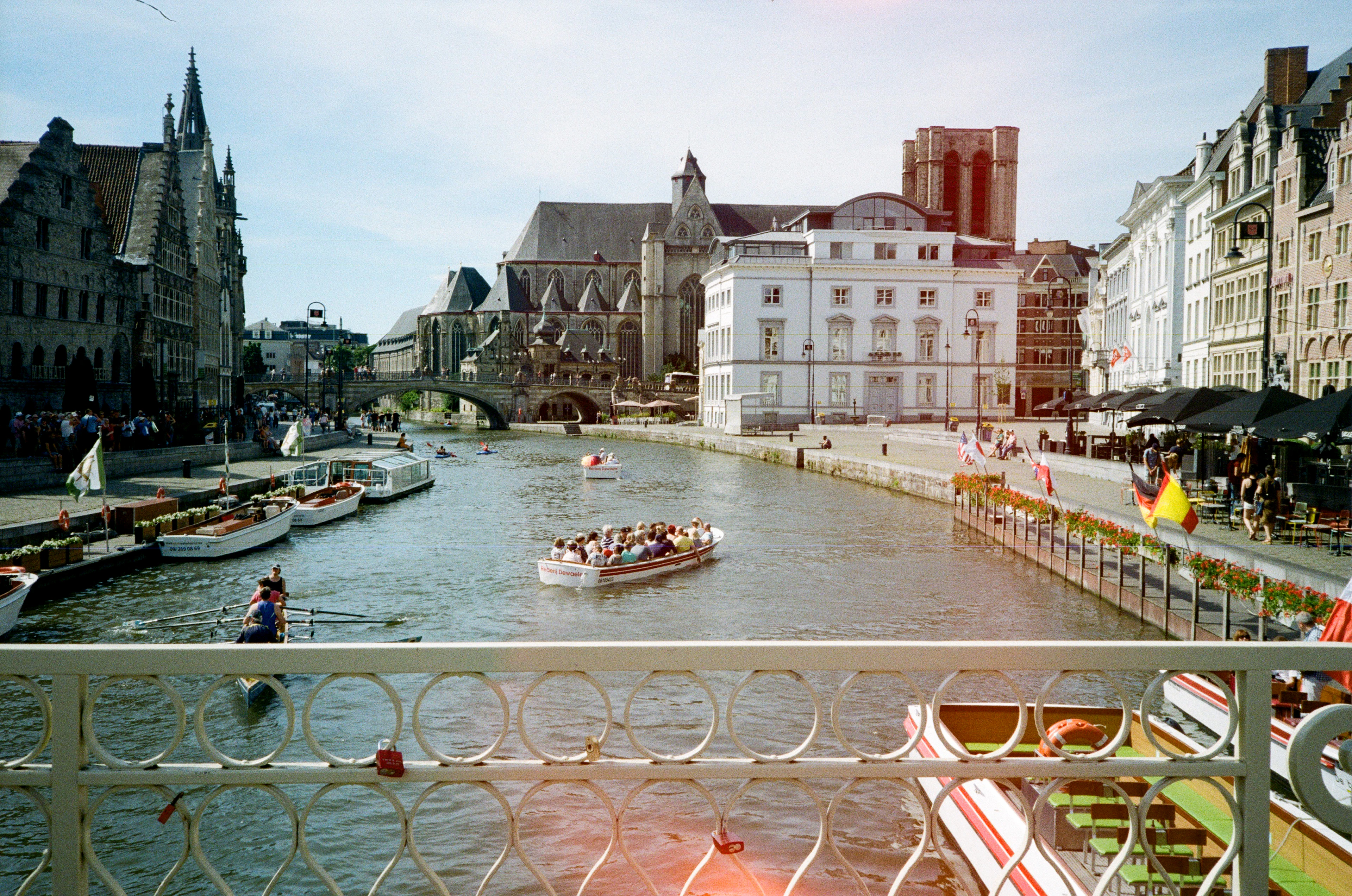 Sunlit Ghent canal seen over a wrought-iron bridge railing, tour boats passing gabled guild houses and a distant spire.