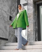 A fashionably dressed person stands on marble stairs. They are wearing a vibrant green blazer, light blue flared jeans, and a white headscarf. The setting appears modern, with large stones and reflective surfaces in the background.