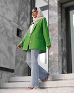 A fashionably dressed person stands on marble stairs. They are wearing a vibrant green blazer, light blue flared jeans, and a white headscarf. The setting appears modern, with large stones and reflective surfaces in the background.