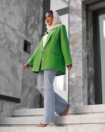 A fashionably dressed person stands on marble stairs. They are wearing a vibrant green blazer, light blue flared jeans, and a white headscarf. The setting appears modern, with large stones and reflective surfaces in the background.