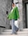 A fashionably dressed person stands on marble stairs. They are wearing a vibrant green blazer, light blue flared jeans, and a white headscarf. The setting appears modern, with large stones and reflective surfaces in the background.