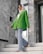 A fashionably dressed person stands on marble stairs. They are wearing a vibrant green blazer, light blue flared jeans, and a white headscarf. The setting appears modern, with large stones and reflective surfaces in the background.