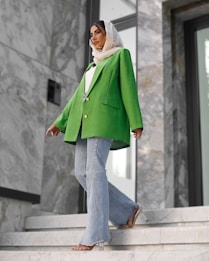 A fashionably dressed person stands on marble stairs. They are wearing a vibrant green blazer, light blue flared jeans, and a white headscarf. The setting appears modern, with large stones and reflective surfaces in the background.
