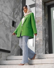 A fashionably dressed person stands on marble stairs. They are wearing a vibrant green blazer, light blue flared jeans, and a white headscarf. The setting appears modern, with large stones and reflective surfaces in the background.