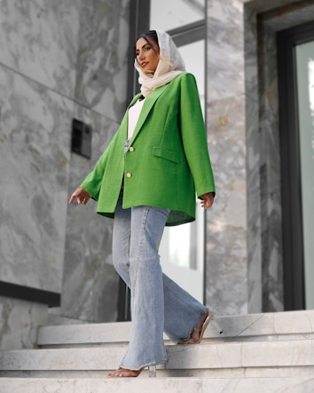 A fashionably dressed person stands on marble stairs. They are wearing a vibrant green blazer, light blue flared jeans, and a white headscarf. The setting appears modern, with large stones and reflective surfaces in the background.